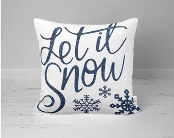 Może przedstawiać: Biała poduszka z napisem "Let it Snow" w niebieskich literach z brokatem. Poduszka ma wzór śnieżynki.