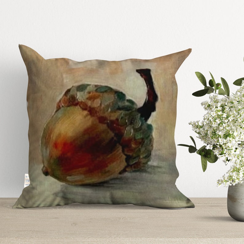 A throw pillow with a painting of a brown acorn with a green cap on a beige background.