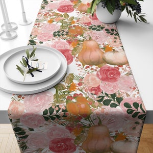 May include: A white table runner with a floral pattern featuring pink roses, green leaves, and orange pumpkins. The runner is perfect for a fall or Thanksgiving table setting.