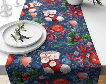 May include: A festive Christmas table runner with a navy blue background. The runner features a repeating pattern of red Christmas stockings, hearts, bows, and holly sprigs. White snowflakes and other festive elements complete the design.