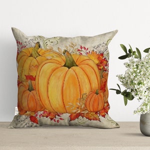 A decorative throw pillow with a fall theme featuring a group of orange pumpkins with green stems and brown leaves on a beige background.