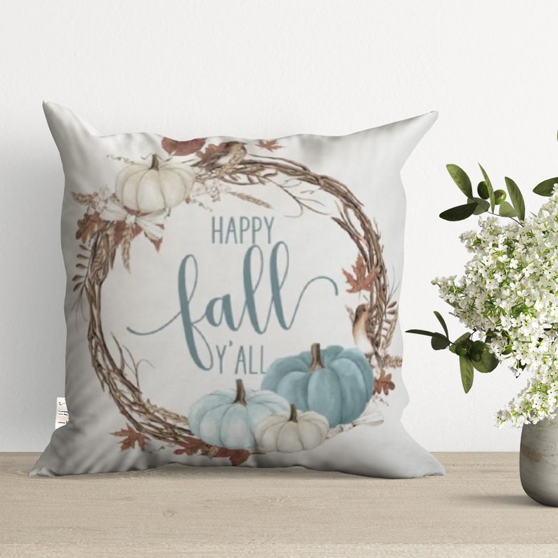 White throw pillow with a blue and brown wreath design. The wreath has pumpkins, leaves, and birds. The text "Happy Fall Y'all" is in the center of the wreath.
