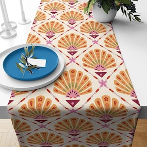May include: A table runner featuring a repeating pattern of orange and yellow fan-shaped motifs with small pink flowers and green foliage accents. The design is set against a cream background with a diamond grid of thin red lines and small pink stars.