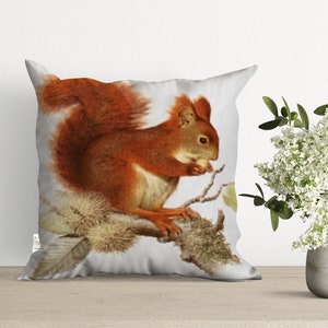 A white throw pillow with a red squirrel eating a nut on a branch. The squirrel has a bushy tail and is sitting on a branch with leaves.