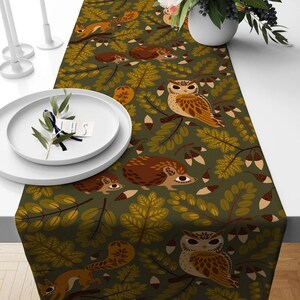 May include: A table runner with a woodland design, featuring owls, squirrels, and hedgehogs amongst oak leaves and acorns. The colour scheme includes browns, greens, and tans on a dark green backdrop. The runner is designed to decorate a table.