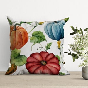 A white pillow with a watercolor design of pumpkins, leaves, and vines. The pumpkins are orange, red, and blue.