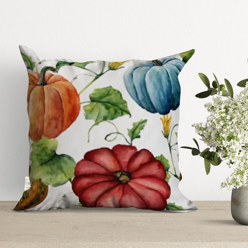 A white pillow with a watercolor design of pumpkins, leaves, and vines. The pumpkins are orange, red, and blue.