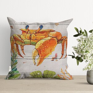 May include: A decorative throw pillow with a watercolor illustration of a crab on a wood plank background. The crab is orange and yellow with blue eyes. The background is white with green and orange accents.