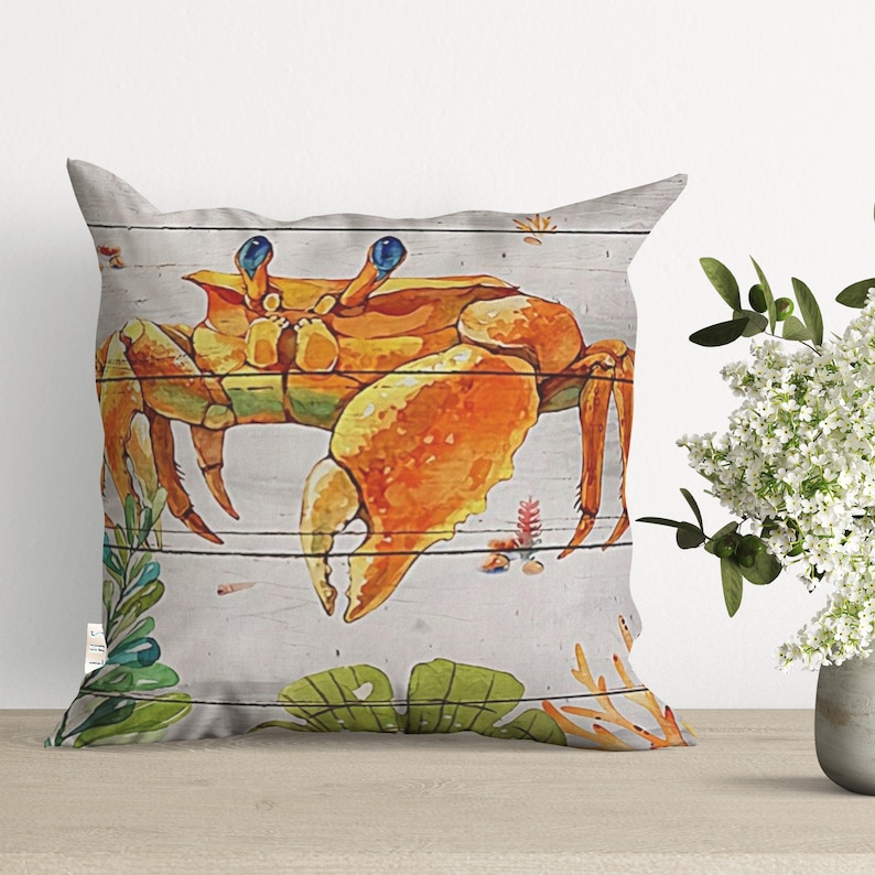 May include: A decorative throw pillow with a watercolor illustration of a crab on a wood plank background. The crab is orange and yellow with blue eyes. The background is white with green and orange accents.