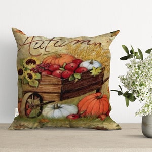 A decorative pillow with a vintage autumn theme. The pillow features a wooden wagon filled with pumpkins, apples, and sunflowers. The text "Autumn" is written in a decorative font on the pillow.