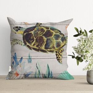 May include: A decorative throw pillow with a watercolor design of a sea turtle swimming in the ocean. The background is a light gray with a wood grain pattern. The turtle is green, brown, and yellow with a black shell. There are also coral, seaweed, and shells in the design.
