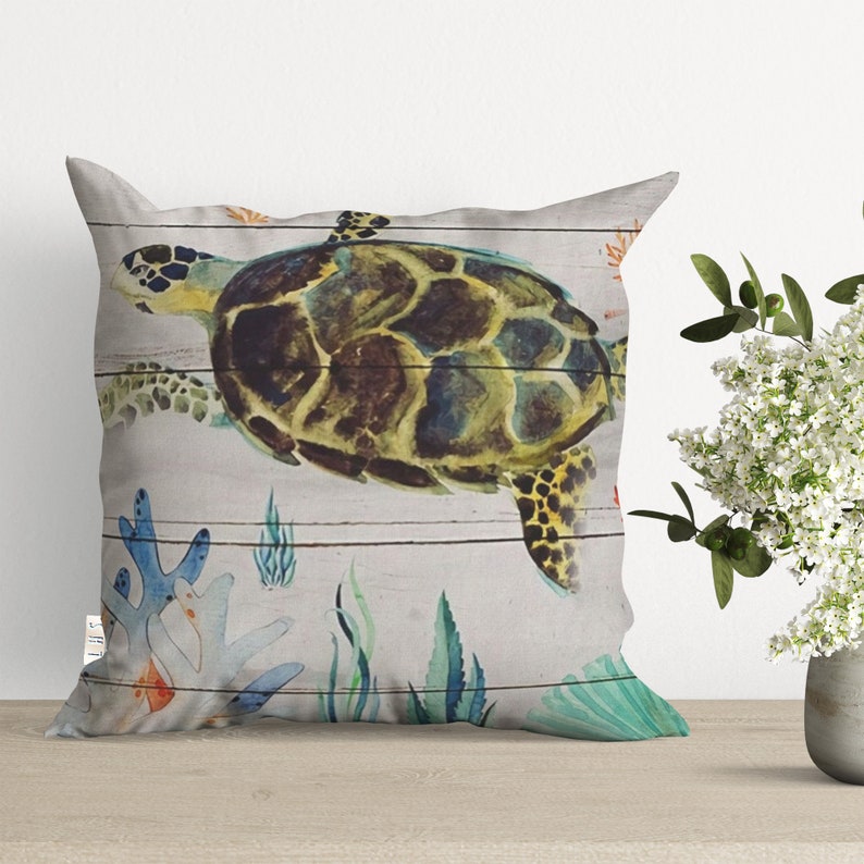 May include: A decorative throw pillow with a watercolor design of a sea turtle swimming in the ocean. The background is a light gray with a wood grain pattern. The turtle is green, brown, and yellow with a black shell. There are also coral, seaweed, and shells in the design.