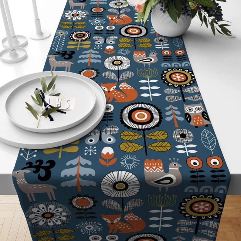 May include: A blue table runner featuring a forest-themed design. The pattern includes foxes, deer, owls, birds, and flowers in orange, white, and grey. The runner is on a white table, with white plates and candles.