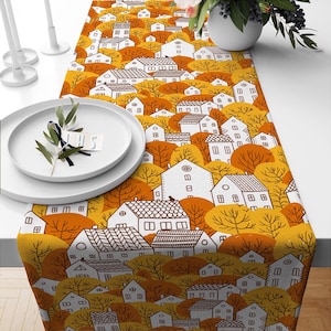Könnte beinhalten: Ein gelber und orangefarbener Tischläufer mit einer weißen und braunen Illustration einer Stadt mit Häusern und Bäumen. Der Läufer liegt auf einem weißen Tisch mit weißen Tellern.