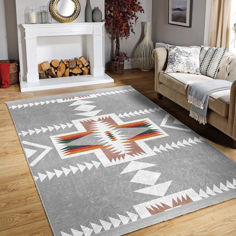 May include: A gray area rug with a white geometric pattern. The pattern features a large cross shape with smaller triangles and diamonds. The rug has a fringe border.