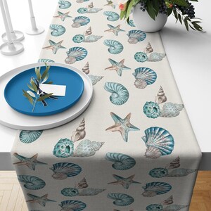 May include: A table runner featuring a repeating pattern of watercolor-style seashells, starfish, and sea urchins in shades of blue, teal, and beige on a light cream background. The runner is displayed with a blue plate and white dinnerware.
