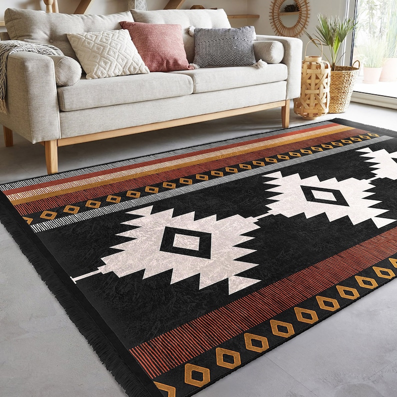 May include: Black and white geometric patterned rug with red and gold accents. The rug has a fringe border and is laid out on a grey floor in front of a light grey sofa.