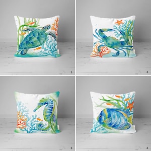 May include: Four decorative pillow covers with watercolor illustrations of sea life. The first pillow features a green sea turtle swimming in blue water with orange coral. The second pillow features a blue crab with orange coral. The third pillow features a blue seahorse with orange coral. The fourth pillow features a blue angelfish with orange coral.