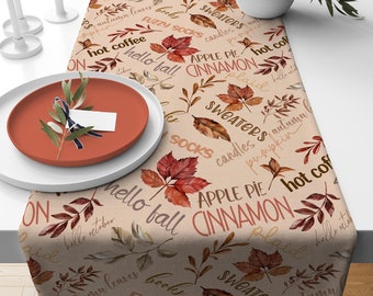 May include: A beige table runner with a fall-themed design. The runner features a repeating pattern of red and orange leaves, along with the words "hot coffee", "hello fall", "apple pie cinnamon", "sweaters", "fuzzy socks", and "pumpkin".