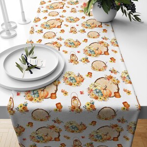 Könnte beinhalten: Weißer Tischläufer mit einem sich wiederholenden Muster aus Kürbissen, Blumen, Laternen und Holzscheiben. Das Muster ist in Orange-, Gelb-, Braun- und Grüntönen gehalten.