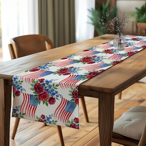 May include: A table runner featuring a repeating pattern of American flags with watercolour-style red roses and small blue flowers on a white background. The design includes flowing flag stripes and star-filled blue sections interspersed with floral clusters.