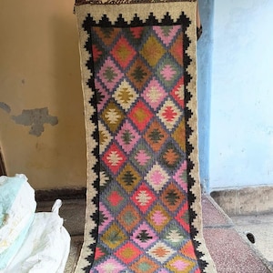 May include: A long, narrow rug with a geometric pattern in shades of pink, blue, green, and brown. The rug has a black border and is made of wool.