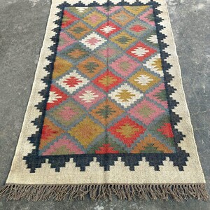 Puede incluir: Una alfombra colorida con un patr&oacute;n geom&eacute;trico, un borde negro y flecos. La alfombra est&aacute; hecha de fibras naturales y tiene un aspecto r&uacute;stico y tejido a mano.