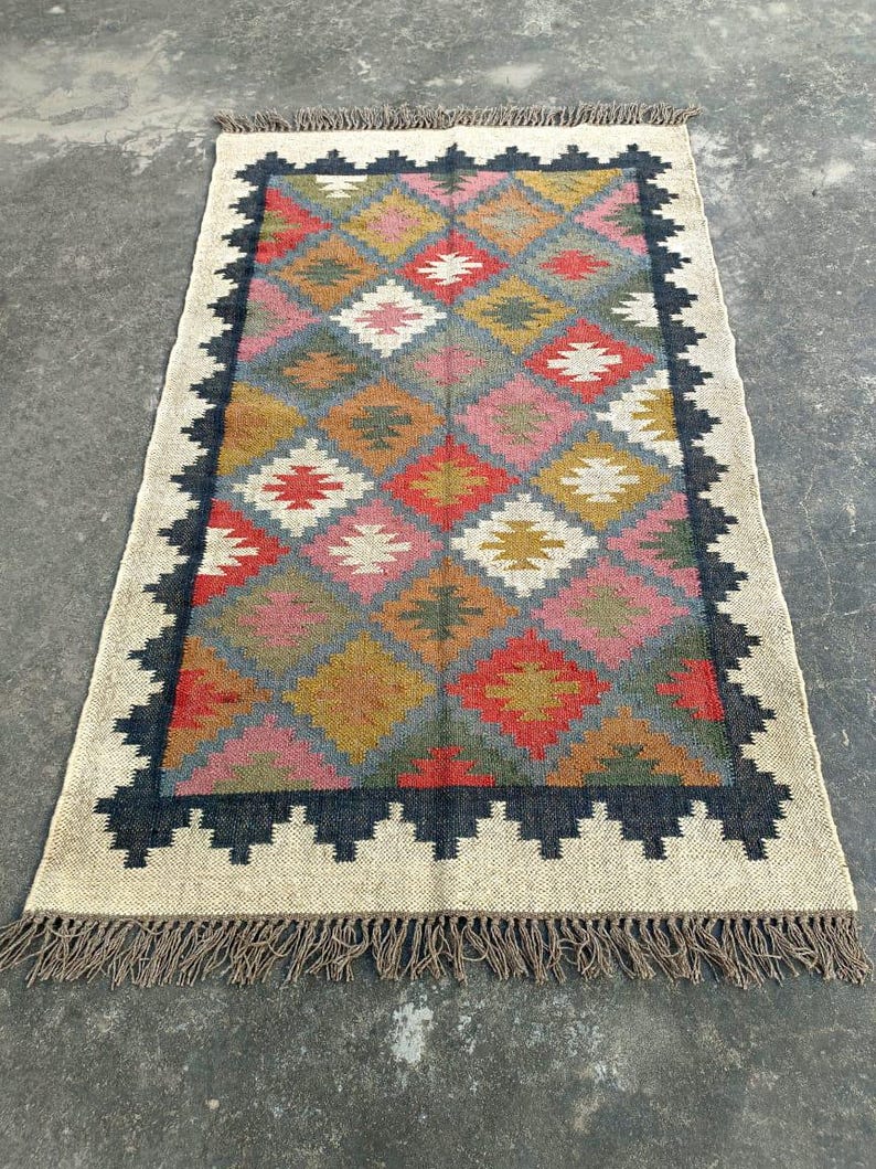 Puede incluir: Una alfombra colorida con un patr&oacute;n geom&eacute;trico, un borde negro y flecos. La alfombra est&aacute; hecha de fibras naturales y tiene un aspecto r&uacute;stico y tejido a mano.