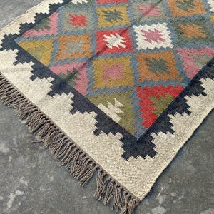 Puede incluir: Una alfombra tejida a mano con un patr&oacute;n geom&eacute;trico en tonos de rojo, azul, verde y marr&oacute;n. La alfombra tiene un fondo blanco y un borde negro con flecos.