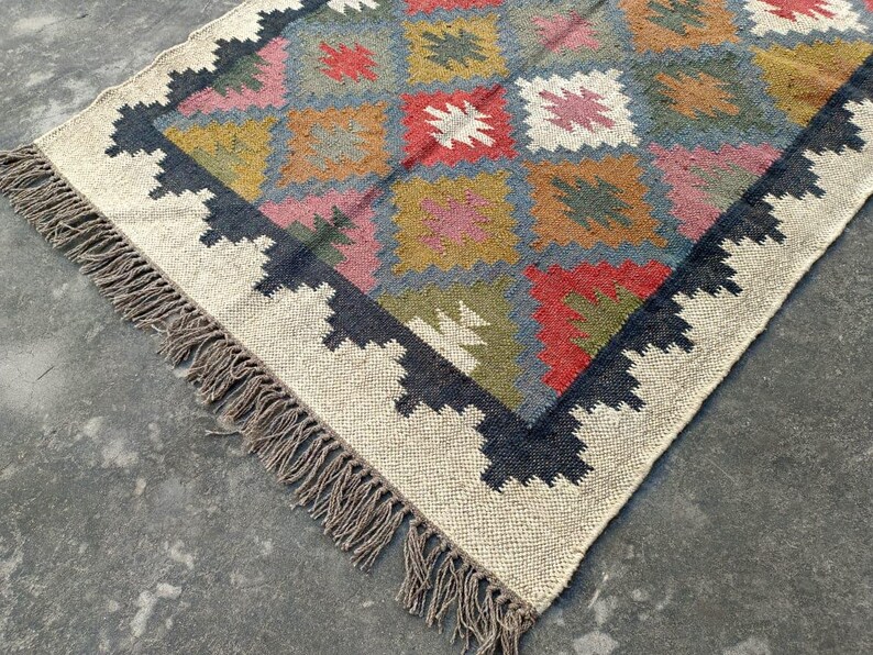 Puede incluir: Una alfombra tejida a mano con un patr&oacute;n geom&eacute;trico en tonos de rojo, azul, verde y marr&oacute;n. La alfombra tiene un fondo blanco y un borde negro con flecos.