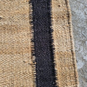 May include: A natural fiber rug with a black stripe down the center. The rug is woven with a textured pattern.