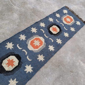May include: A blue and white rug with a geometric pattern of stars and crescent moons. The rug has a fringe border and is made of a woven material.