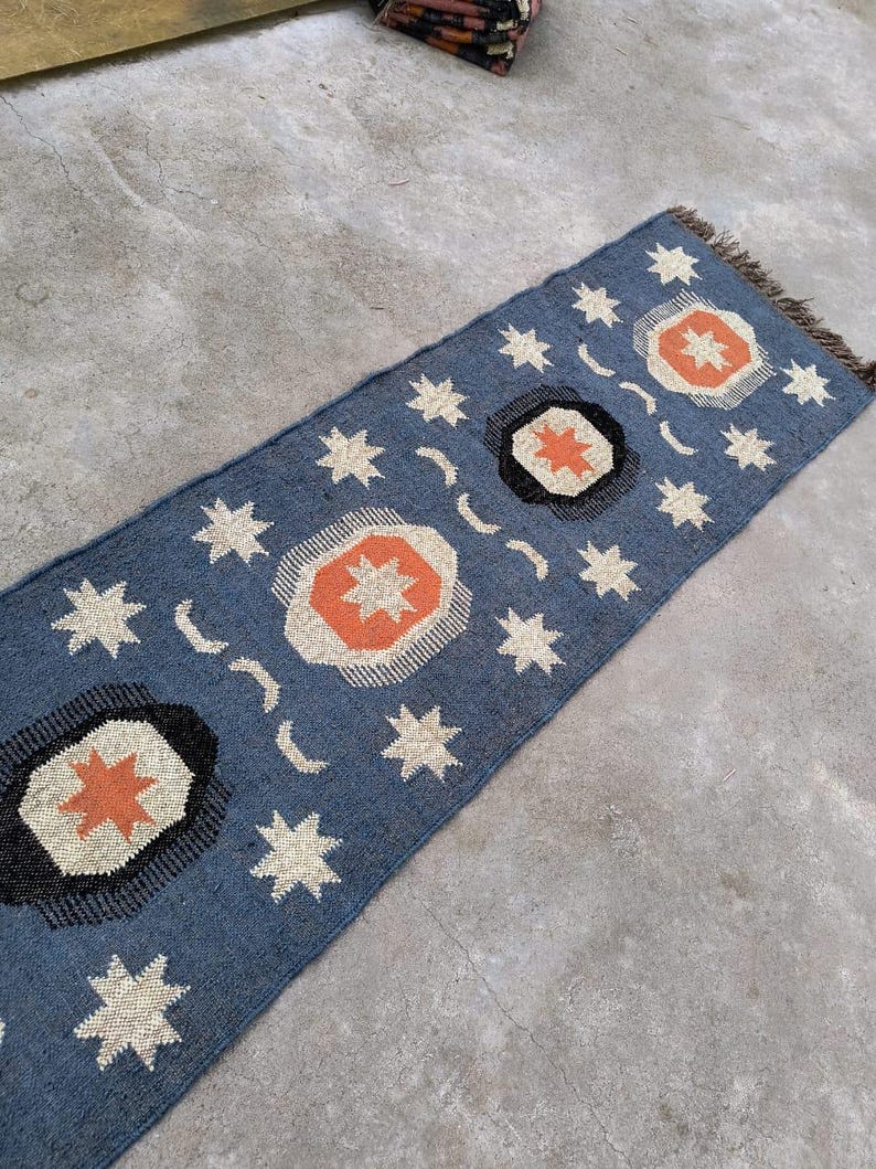May include: A blue and white rug with a geometric pattern of stars and crescent moons. The rug has a fringe border and is made of a woven material.