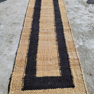 May include: A natural fiber rug with a black border. The rug is woven with a simple pattern and has a natural, earthy look.