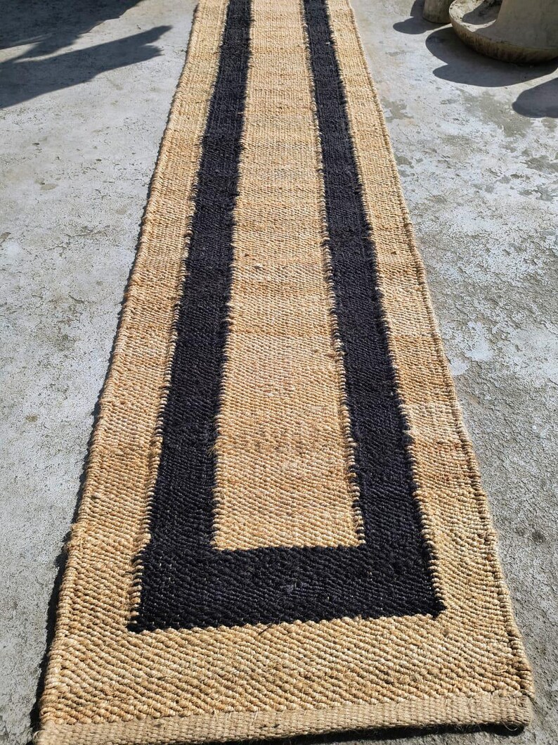 May include: A natural fiber rug with a black border. The rug is woven with a simple pattern and has a natural, earthy look.