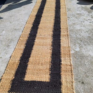 May include: A natural fiber rug with a black and tan striped pattern. The rug is woven with a thick, textured material and has a long, rectangular shape.