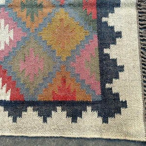 Puede incluir: Una alfombra colorida con un patr&oacute;n geom&eacute;trico en tonos de rojo, rosa, azul, marr&oacute;n y blanco. La alfombra tiene un borde negro y flecos en los bordes.