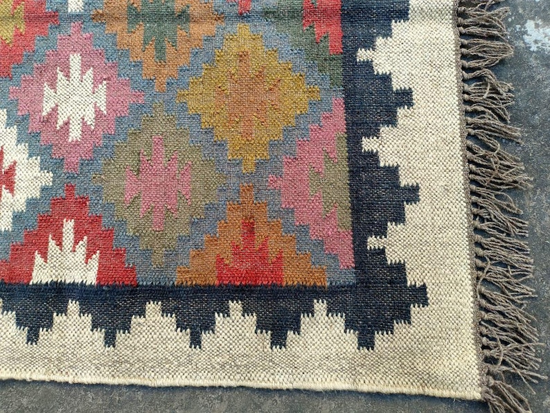 Puede incluir: Una alfombra colorida con un patr&oacute;n geom&eacute;trico en tonos de rojo, rosa, azul, marr&oacute;n y blanco. La alfombra tiene un borde negro y flecos en los bordes.