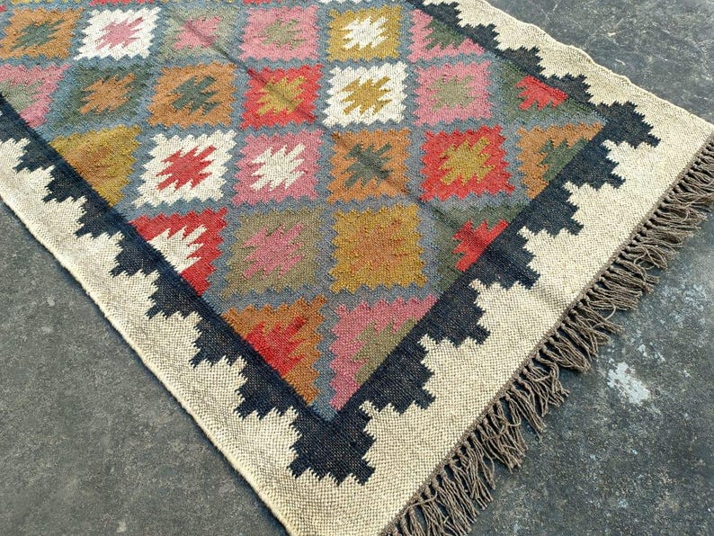 Puede incluir: Una alfombra multicolor con un patr&oacute;n geom&eacute;trico de diamantes y cuadrados. La alfombra tiene un fondo blanco con acentos negros, marrones, rojos, rosas y amarillos. La alfombra tiene un borde de flecos.