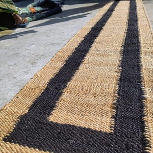 May include: A natural fiber rug with a woven design. The rug has two black stripes running down the center, creating a geometric pattern. The rug is made of natural materials and has a rustic look.