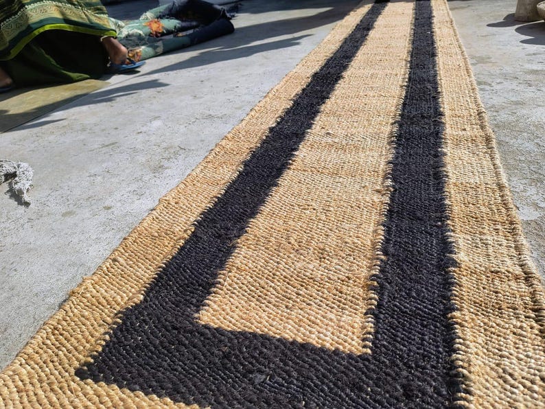 May include: A natural fiber rug with a woven design. The rug has two black stripes running down the center, creating a geometric pattern. The rug is made of natural materials and has a rustic look.