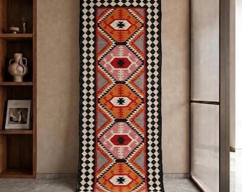 May include: A long, rectangular woven runner rug featuring a repeating geometric pattern of diamonds in orange, pink, and black on a gray background. The rug is framed by a black and white checkered border and finished with fringe at both ends.