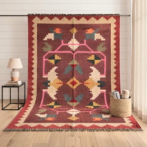 May include: A rectangular area rug featuring a deep maroon background with a geometric pattern in cream, pink, yellow, blue, and orange. The rug has a wide cream-coloured border with a zigzag edge and short fringe along the top and bottom ends.