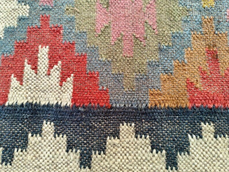 Puede incluir: Primer plano de una alfombra tejida con un patr&oacute;n geom&eacute;trico en tonos rojos, azules, marrones y blancos. La alfombra tiene una superficie texturizada y un borde de flecos.