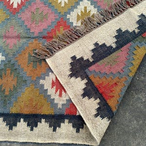 Puede incluir: Una alfombra tejida a mano con un patr&oacute;n geom&eacute;trico en tonos de azul, marr&oacute;n, rojo y blanco. La alfombra tiene un borde de flecos y est&aacute; hecha de fibras naturales.