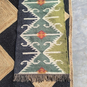 May include: A rectangular, patterned rug with a geometric design in shades of blue, green, and beige. The rug has a fringed edge and is placed on top of a larger rug with a black and beige diamond pattern. The rug is suitable for home decor.