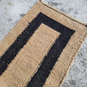May include: A natural fiber rug with a black border. The rug is made of woven straw or sisal and has a rectangular shape.