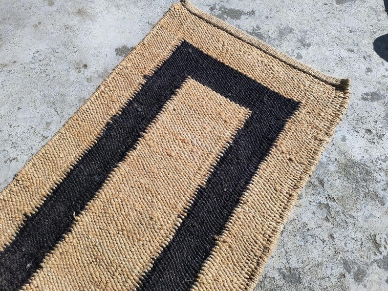 May include: A natural fiber rug with a black border. The rug is made of woven straw or sisal and has a rectangular shape.