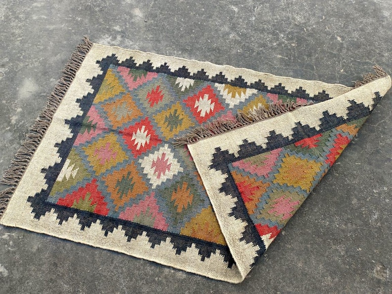 Puede incluir: Una alfombra colorida con un patr&oacute;n geom&eacute;trico en tonos de rojo, amarillo, azul, verde y marr&oacute;n. La alfombra tiene un borde de flecos y est&aacute; hecha de lana.