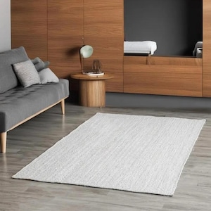 Puede incluir: Una alfombra blanca tejida con una textura sutil, colocada sobre un suelo de madera clara en un salón. La alfombra está delante de un sofá gris y una mesa auxiliar de madera con una lámpara.